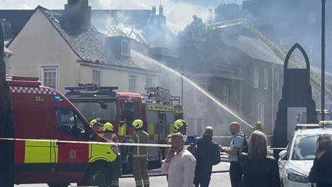 Firefighters at the scene of a house fire in a street. Crews in yellow hardhats and brown overalls work with each other. A firefighter in the distance directs a water hose into a home. 