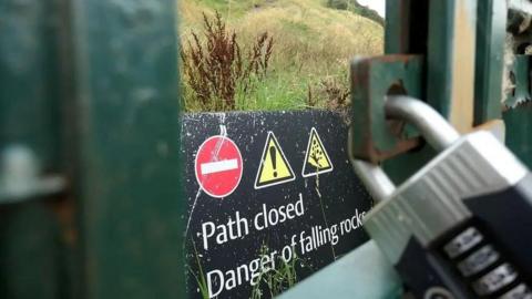 A close up photo of a padlock on a gate at the Radical Road, and a warning sign that reads: "Path closed. Danger of falling rocks". Behind the gate a section of the path is just visible.