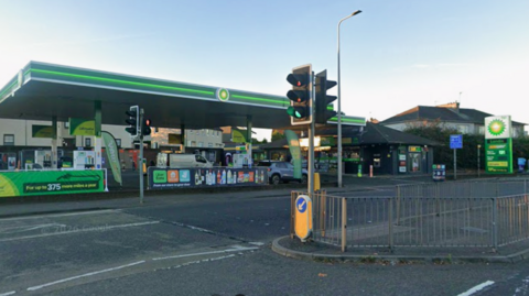 Shell petrol station on roadside. It is mostly green and there are a set of traffic lights in the foreground