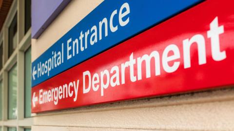 An image of a hospital sign showing the main entrance and the emergency department. The signs are blue and red and have arrows.