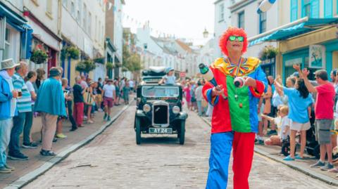 A view of a parade, a person dressed as a clown is juggling, while a child rides in a vintage car behind her. People are lining the streets.