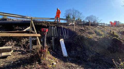 A view from halfway up the landslip at Tinsley Green. A Network rail worker is wearing orange high-viz trousers and jacket and a number of repair tools can be seen near the affected area.