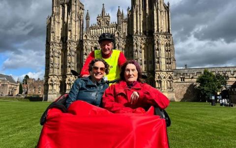 Two women say in a trishaw, covered in a red blanket with a man in high-vis and a helmet steering behind them. They are outside Wells Cathedral 