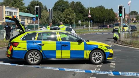 A police car with its boot open is parked across a road behind blue and white police tape. Police officers are talking to each other and inspecting the scene, by a pedestrian crossing with traffic lights. Other vehicles can be seen parked in the background.