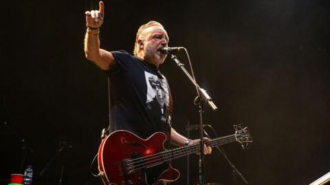  Peter Hook playing a red bass and singing into a microphone- he wears a black T-shirt with Oasis on the front - he's performing at Wembley arena