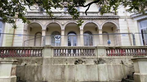 A historic building with a balcony with arches and columns. The cream coloured building shows signs of disrepair. A metal fence is blocking access to the building. Tree branches can be seen above.