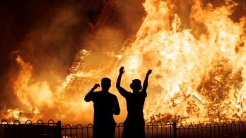 Two men stand in front of a bonfire, silhouetted by its flames. There is a metal fence in front of them and one is wearing a bucket hat. 