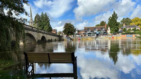 Water can be seen under a bench next to a river that has burst its banks, with a black and white building 