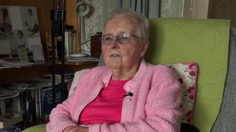 Gloria Phillips in a pink cardigan, sat in an armchair. She has short, grey hair and glasses.