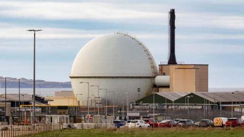 The large sphere-shaped structure housing a nuclear reactor is sited next a box-shaped building with a chimney. There are other buildings and vehicles parked in a car park.