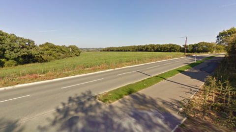 Streetview image of Stag Lane - a straight road with fields and trees behind and a wide footpath in the foreground