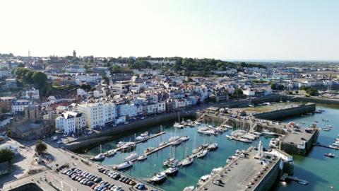 Aerial view of a harbour. Boats are moored in the water. Cars are parked in a car park next to the harbour. Properties are built up next to the water. Properties can be seen in the distance.