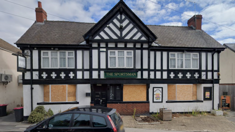 The pub features a mock-Tudor architectural style, with black timber framing against a white plastered exterior and a grey slate roof with two chimneys.