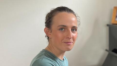 Amy Symington has brown hair which is tied back off her face. She is wearing a green t-shirt. Her face is towards the camera, looking over her right shoulder. In front of her slightly out of focus is the edge of an upright piano. 