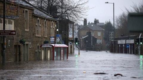 A street is submerged under flood water. There is a traffic light on green sticking out from the water in the centre of the picture, as well as the top half of a bus stop. Brick buildings on either side of the street are also submerged in water up to their bottom floor windows.