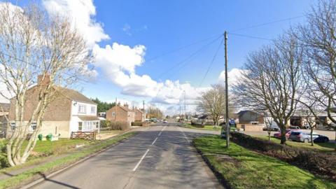 A Google street view of an empty road with a car park on the right and a few detached houses on the left.