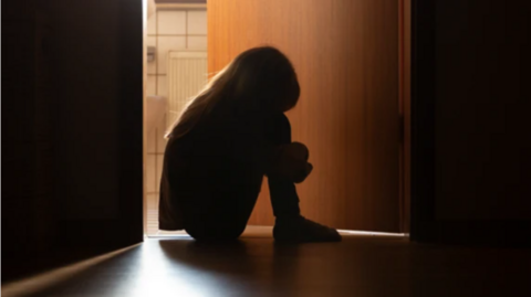 The silhouette of a child sitting in the dark on the floor with their head bowed down and their knees tucked in to their chest, with a door and a crack of light showing another room in the background.