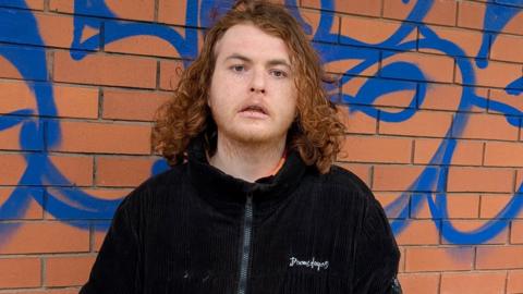 Theo Godfrey-Cass standing in front of an external wall of the gallery. The brick wall has been sprayed with a graffiti tag in blue paint. Mr Godfrey-Cass has shoulder-length red hair and is wearing a black coat. He is very unhappy.