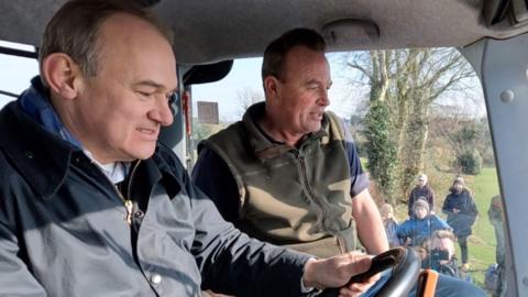 Sir Ed Davey, dressed in a navy blue wax jacket, steers a tractor sat next to Chris Blaxell, wearing a zipped-up green gillet. Photographers are seen in the background through the tractor window.