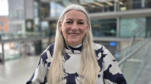 Kay Mason Billig is standing on a balcony in a glass-fronted building. She is wearing a blue and white patterned long-sleeve top.
