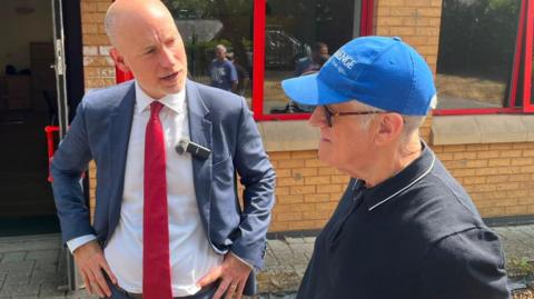 Stephen Kinnock wearing a suit and red tie stands outside a brick community centre, speaking with a member of a local men's wellbeing group who is wearing a blue baseball cap and dark polo shirt. They are beside a building's entrance, which has red-framed windows and an open door. Bright sunshine casts shadows across the paved area outside the centre.