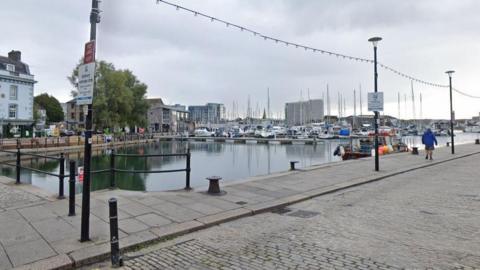 Google Street View image of the Barbican area of Plymouth. A harbour with several yachts moored up is in the centre of the area. A man wearing shorts and a blue coat is walking along the water's edge on the pavement.