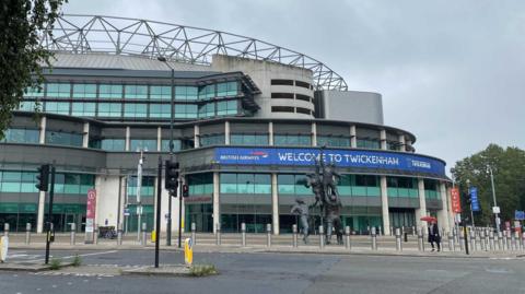 Image shows the front entrance of Twickenham stadium. There is a blue banner that displays welcome to Twickenham and has a British Airways logo on the left. There is a bronze statue of a rugby line-out in front of the stadium. There are traffic lights and a pedestrian crossing visible on the left of the image. 