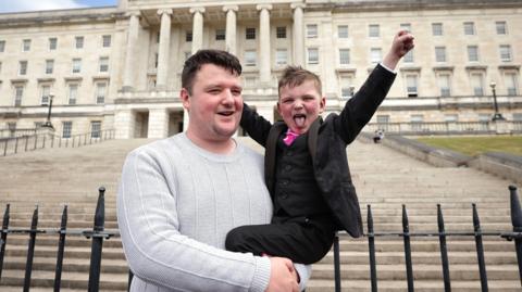 A young boy in his father's arms with his arms in the air and sticking his tongue out. They are standing in front of Parliament buildings at Stormont.