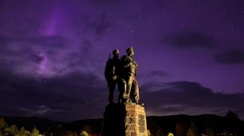 The aurora borealis glows purple behind the Commando Memorial at Spean Bridge. Two of the figures on the memorial are visible. The figures are portrayed wearing military fatigues.