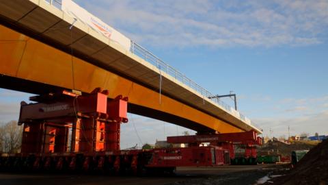 A view of the new bridge, from the M6 northbound carriageway, as it is moved into place. The large steel bridge is brown and white. It is being rolled into place across the motorway on red steel machinery.