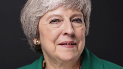 Theresa May, former UK prime minister, with short grey hair, wearing a green top against a dark background