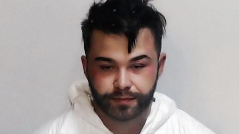 Mugshot of a man in a white forensic-style protective suit with a hood, standing against a plain light gray background. The person has short, dark, slightly messy hair with small white specks visible in the hair