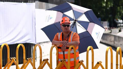 A worker in a high vis orange vest and helmet stands in the shade with a parasol above their head while standing next to a yellow barrier on a closed footpath in London on Tuesday - there is a sign next to him reading 'pedestrians please use other footpath'
