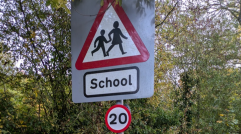 A school road sign that depicts two people within a red triangle and includes the word School. A 20mph sign is below that and trees are in the background.