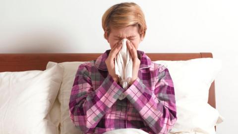 A person sneezing into a tissue, she is wearing a pink and purple check pyjama top. She has light hair.