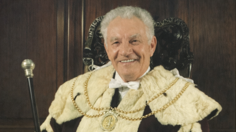 A smiling grey-haired man in robes with a fur-lined collar and a ceremonial chain on top. He is wearing a white bow tie and carrying a mace. There is a panelled wood background behind him.