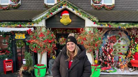 A woman with long brown hair wearing a black coat and black beanie. She is standing in front of a house which is decorated with a Grinch Christmas display.