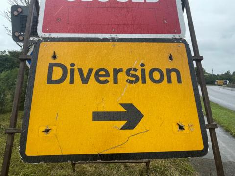 A bright yellow road sign which says "Diversion" in black with a black arrow underneath. It is resting on an verge, with a road on the right. In the far distance beyond the sign is a lorry.