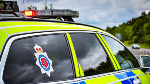 A police car is parked facing a road. It has a sticker on the back window which reads Lancashire Constabulary. A blue sky and some trees can be seen in the distance over a road. 