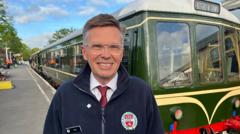 A man in glasses and a blue fleece stands besides a green train with cream and red markings. 