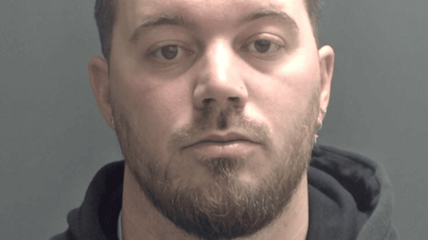 Christopher Rudd is staring at the camera and has a light brown and beard and earrings. He is wearing a dark hoodie and is standing against a plain grey background.