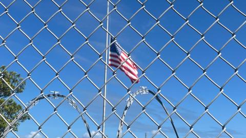 An American flag flies against a blue sky behind a chainlink fence and barbed wire at Guantanamo Bay