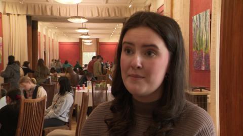 A girl with long thick brown hair and a brown top. She has a neutral expression and stands in front of the camera. Behind her tables with groups of people sitting at them can be seen