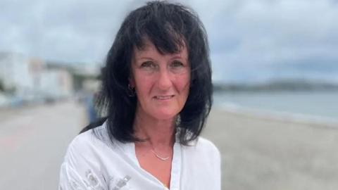 Jackie Hallam, who has shoulder-length black hair, is wearing a white open-necked blouse. She is standing on Douglas Promenade, with the sandy beach and shoreline on the right and Douglas Promenade on the left.