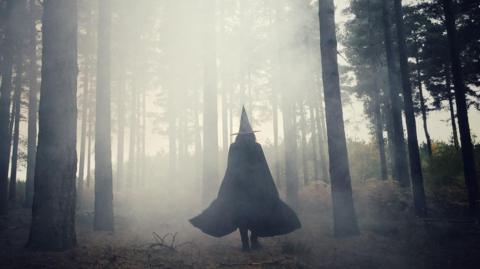 A woman with black cloak and a hat, walking through a misty forest