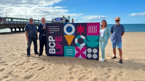 Three men and one woman posing for a picture on a beach with a big BCP Life placard for the BCP Life card.