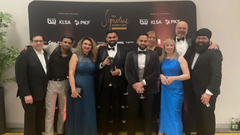 Nine men and women wearing various forms of formal evening dress pose for a picture at an awards ceremony. The man in the middle, in a dinner jacket and bow tie, is holding a black trophy. The group is standing in front of a black branded media wall featuring several sponsor names alongside the event name, The Signature Awards 2026.  