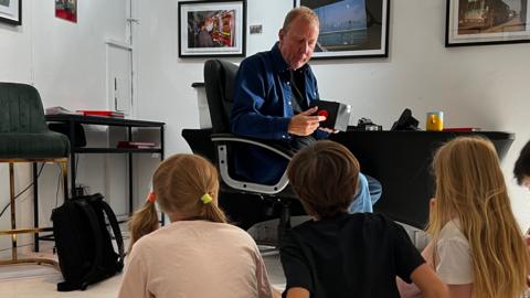 The image shows a man sat on a chair talking to a group of five children who are all sat on the floor. Framed pictures can be seen on the wall in the background.