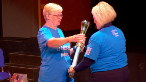 The image shows a ceremonial moment where two individuals in blue shirts are participating in a symbolic torch handover. One person is passing the torch with both hands, while the other is receiving it with one hand. The setting appears to be indoors, possibly on a stage, with chairs and equipment in the background.