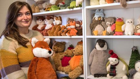 Amanda Hope stands holding a toy fox next to her collection of Jellycat toys stacked on shelves